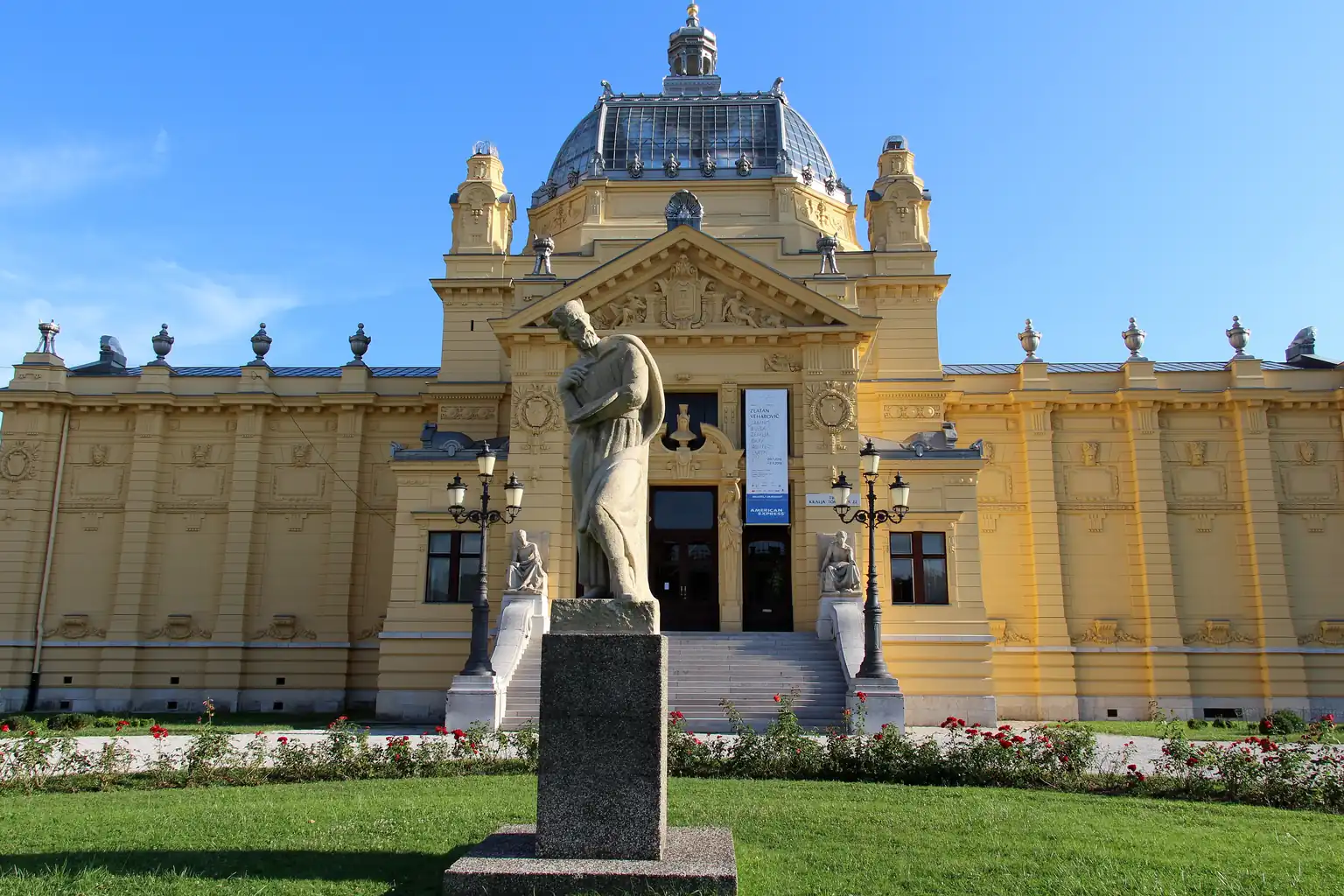 Front of the yellow Art Pavilion in Zagreb, with a stone statue and red roses on the lawn.