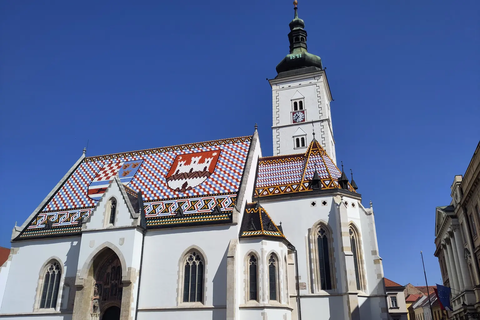 St Mark's Church in Zagreb, its tiled roof showing the Croatian and city coats of arms, under blue sky.