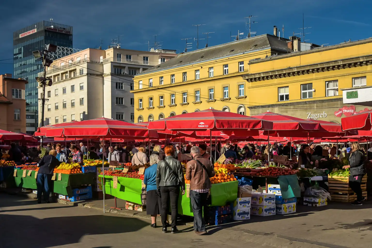 Dolac market in Zagreb, with red umbrellas over fruit and vegetable stalls and shoppers in front of yellow buildings.